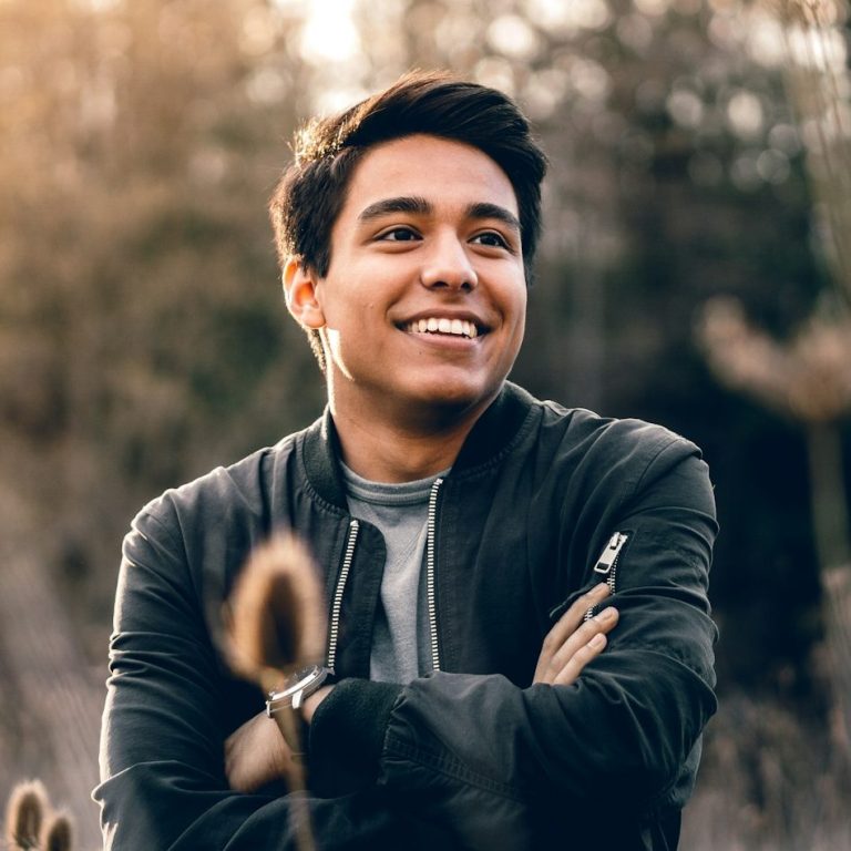 Smiling young man in a black jacket, standing in a grassy field with blurred trees behind him.